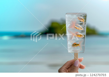Transparent fruit ice cream with small golden fish frozen in ice cream, surreal dessert, female hand holding delicious unique dessert on background of beach with clear blue sky 124270703