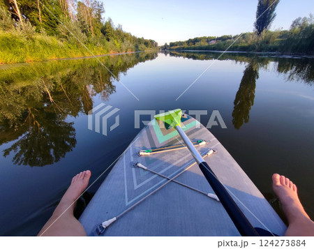Female feet and SUP board with green paddle on surface of river water with trees on banks on calm windless summer evening. Person swimming on SUP board on pond with reflective water surface Female feet and SUP board with green paddle on surface of river water with trees on banks on calm windless summer evening. Person swimming on SUP board on pond with reflective water surface 124273884