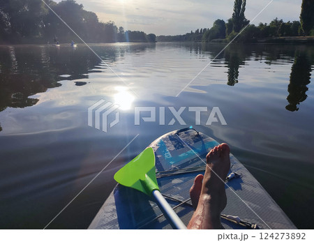 Bare legs of woman on SUP board on surface of river or lake with sun reflecting and with trees on banks on warm summer evening. Person swimming on SUP board on pond on calm day. Tourism rest sport 124273892