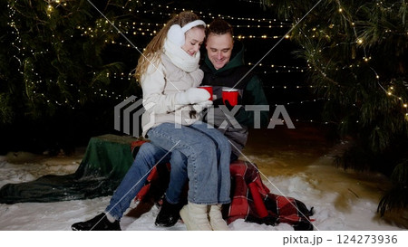 Romantic couple cuddling on bench, drinking warm beverages amid glowing christmas market lights, sharing intimate moment surrounded by festive winter ambiance 124273936