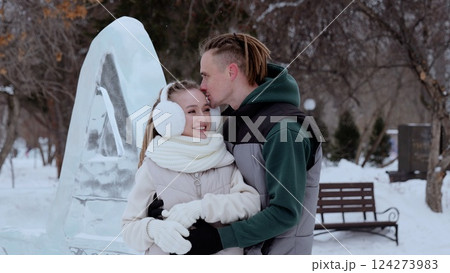 Loving couple cuddling near intricate ice sculpture, sharing tender moment amid snowy park landscape, expressing winter romance and intimate connection 124273983