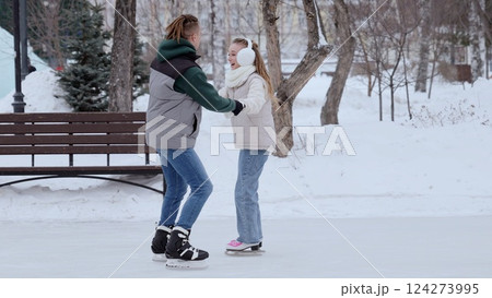 Young couple holding hands while gliding across icy rink, sharing loving moments amid snow covered park landscape, winter romance unfolding with joyful connection 124273995