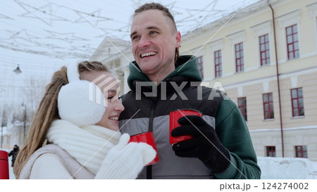 Loving couple sipping warm beverages, snuggling together amid falling snow, embodying winter romance in urban landscape 124274002