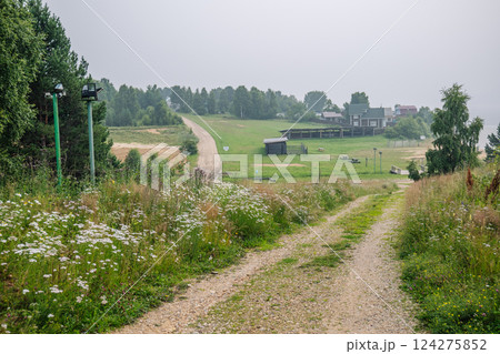Winding gravel path traversing wildflower meadow toward weathered ranch buildings amid misty russian landscape 124275852