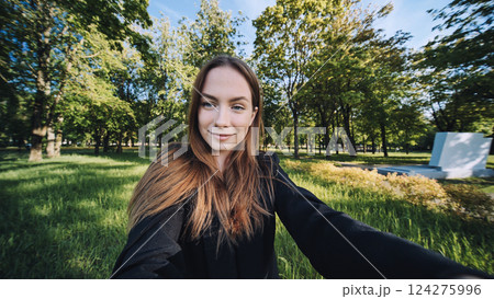 Young woman with flowing brown hair capturing summer moment while taking selfie amid lush green park landscape 124275996