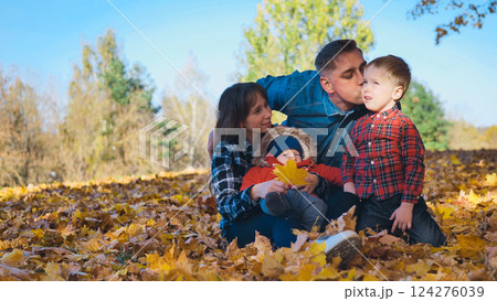 Smiling family sitting on fallen leaves in a park during a beautiful autumn day, father kissing his son 124276039