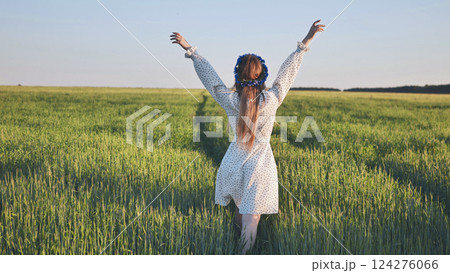 Young woman wearing flower wreath walking in green wheat field with arms outstretched enjoying freedom and nature 124276066