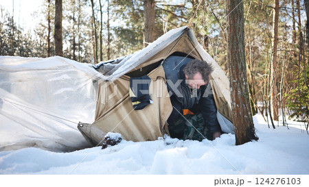 Homeless man crawling out of makeshift tent in a snowy forest during a frigid winter day, facing the harsh realities of survival Homeless man crawling out of makeshift tent in a snowy forest during a frigid winter day, facing the harsh realities of survival 124276103