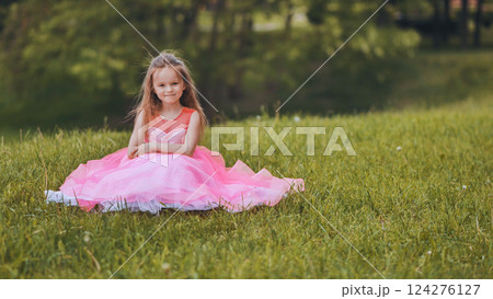 Young girl in pink dress sitting on grassy lawn, basking in warm summer sunlight, smiling happily Young girl in pink dress sitting on grassy lawn, basking in warm summer sunlight, smiling happily 124276127