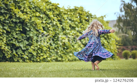 Young blonde girl spinning playfully in white sundress, dancing barefoot on lush green lawn near garden hedgerow, radiating childhood joy 124276206