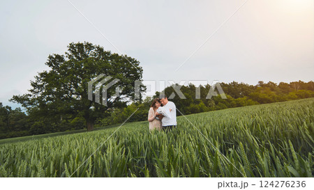 Loving parents holding newborn amid golden wheat, peaceful oak tree backdrop creating serene family landscape at sunset 124276236