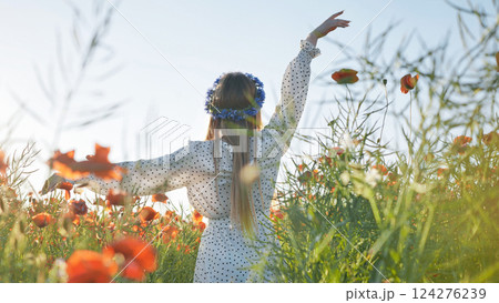 Young woman wearing flower crown, polka dot dress, standing amid blooming poppies with arms spread wide, embracing scenic landscape and natural beauty 124276239