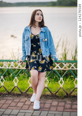 Young woman leaning on lakeside fence, wearing floral dress and denim jacket, basking in golden sunset light Young woman leaning on lakeside fence, wearing floral dress and denim jacket, basking in golden sunset light 124276589