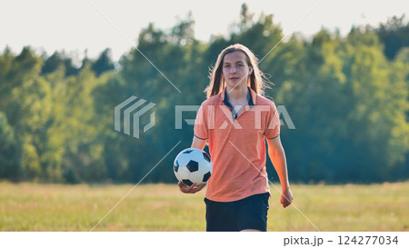 Teenage boy with long hair strolling through a grassy field with a soccer ball after a fun game with friends 124277034