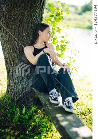 Teenage girl relaxing by lakeside, resting beneath shady tree during golden summer moment Teenage girl relaxing by lakeside, resting beneath shady tree during golden summer moment 124277194