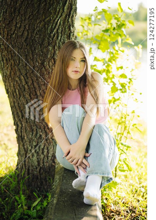 Young woman enjoying summer day in park, sitting under a tree and wearing casual clothing Young woman enjoying summer day in park, sitting under a tree and wearing casual clothing 124277195