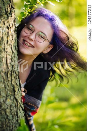 Teenage girl with purple hair, glasses, and braces peeking from behind tree bark during bright summer day 124277213