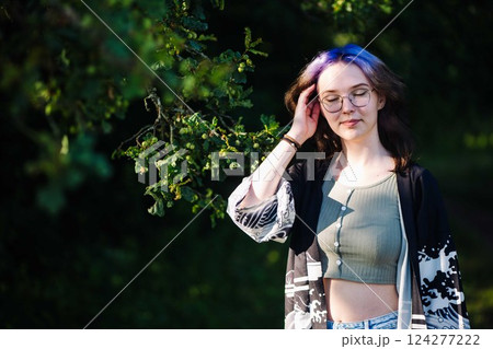 Teenager with purple hair and glasses enjoying a peaceful moment in nature, touching her hair under the shade of a tree 124277222