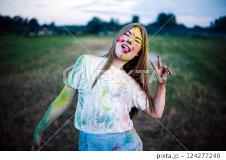 Playful teen girl sticking out her tongue and making victory sign at holi festival, covered in colorful gulal powder 124277240