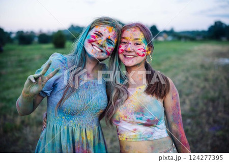 Two young women covered in colorful powder embrace, one making peace sign, enjoying the vibrant atmosphere of holi festival 124277395