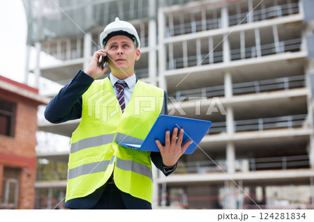 Man architect in helmet with paper documents talking phone at construction site 124281834