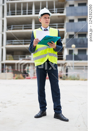 Architect in protective helmet and jacket with folder of documents on construction site 124282260