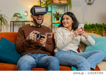 Happy young man wearing VR goggles playing videogame with joystick sitting beside Indian girlfriend Happy young man wearing VR goggles playing videogame with joystick sitting beside Indian girlfriend 124287393