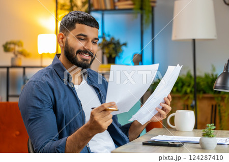 Indian man freelancer working with documents from home office analyzing reading business report Indian man freelancer working with documents from home office analyzing reading business report 124287410