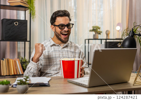 Tired business man taking break from work wearing 3D glasses earing popcorn watching film on laptop Tired business man taking break from work wearing 3D glasses earing popcorn watching film on laptop 124287421