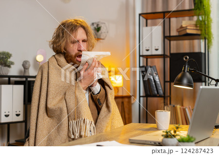 Sick businessman shivering from the cold wrapped in blanket sneezes works on laptop at office 124287423