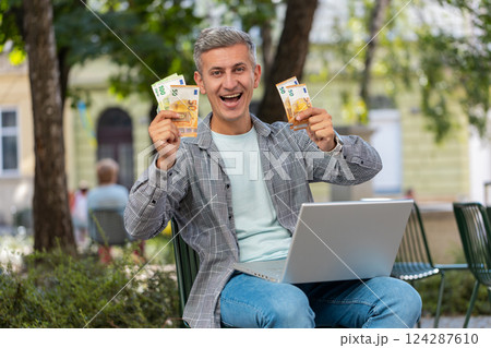 Happy rich man using laptop winner victory holding cash money in euro banknotes sitting on chair Happy rich man using laptop winner victory holding cash money in euro banknotes sitting on chair 124287610