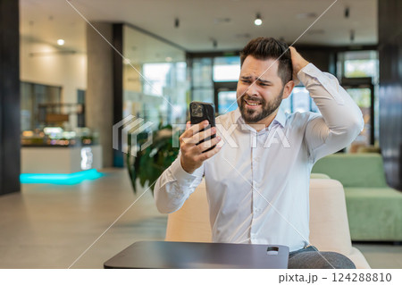 Young businessman using mobile phone becomes shocked sudden lottery results in office hotel lobby Young businessman using mobile phone becomes shocked sudden lottery results in office hotel lobby 124288810