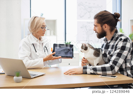 Female veterinarian discussing x-ray image with adult male client holding cat during visit. Professional consultation environment with laptop, medical equipment, and educational posters. Female veterinarian discussing x-ray image with adult male client holding cat during visit. Professional consultation environment with laptop, medical equipment, and educational posters. 124289846