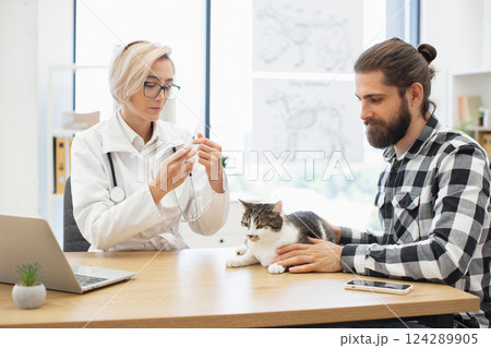 Veterinarian prepares injection with care, while male owner comforts cat during examination. Image shows trust, care, professional service at veterinary clinic. 124289905