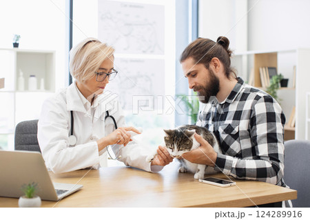 Male owner and female veterinarian discuss care of injured cat in clinic room. Veterinarian treats cat's injury, showing care and expertise, while owner shows concern and care for pet's wellbeing. 124289916