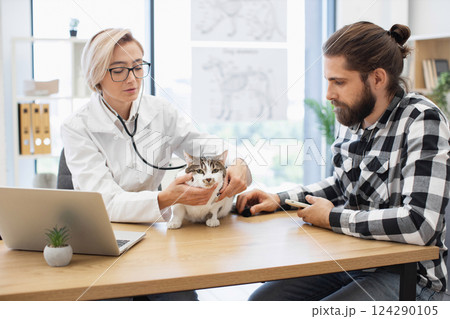 Female veterinarian examining domestic cat at modern veterinary clinic while bearded male owner watches attentively. 124290105