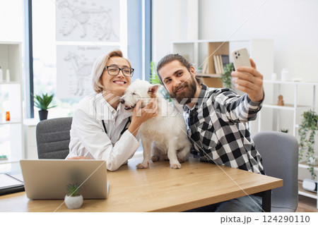 Bearded man capturing selfie with smiling female veterinarian holding small white fluffy dog indoors. White woman approximately late 30s and man creating friendly bond in pet care environment. 124290110