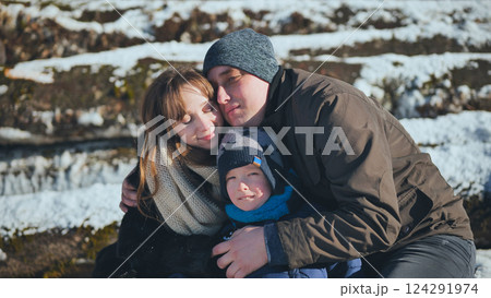 Family warmly hugging during snowy winter walk, surrounded by sunlit trees and pristine white landscape 124291974