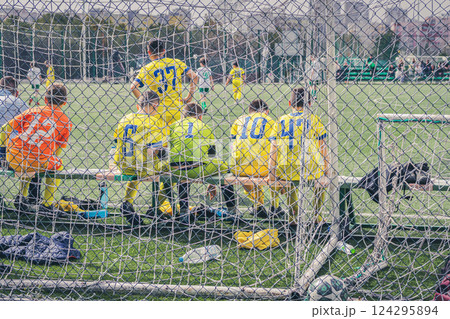 Unrecognizable boys football team in yellow uniform sitting on bench behind goal net on green field. Concept of teamwork, competition and youth sports spirit Unrecognizable boys football team in yellow uniform sitting on bench behind goal net on green field. Concept of teamwork, competition and youth sports spirit 124295894