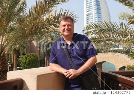 A confident adult man poses for a photo against the stunning backdrop of a famous hotel in Dubai, showcasing the luxurious architecture and vibrant atmosphere of the city, capturing a moment of joy 124302773
