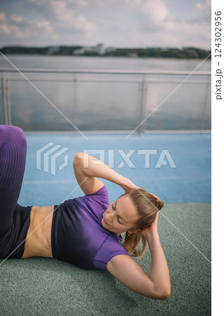 Woman performing bicycle crunches by the waterfront during a bright, cloudy afternoon workout 124302956
