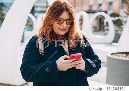 Happy woman chatting on phone and relaxing outdoors. Urban, people concept. Smiling 30s redhead woman text messaging on smart phone at sunny day on the city street, spring time 124308164