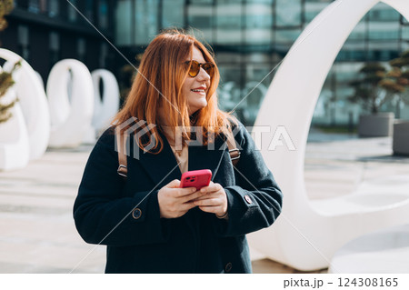 Happy woman chatting on phone and relaxing outdoors. Urban, people concept. Smiling 30s redhead woman text messaging on smart phone at sunny day on the city street, spring time 124308165