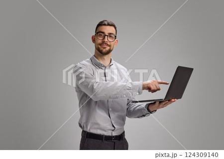 Young man pointing at laptop screen with confident smile, standing in modern office clothes. Young man pointing at laptop screen with confident smile, standing in modern office clothes. 124309140