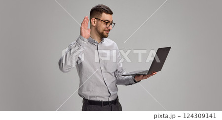 Smiling employee raising hand in front of laptop during video call and waving to colleagues 124309141