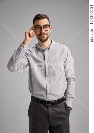 Portrait of young man adjusting glasses, wearing shirt and black pants, looking with confidence and little smile against grey studio background 124309151