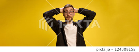 Portrait of shocked and excited young man in black and white outfit holding hands behind head with wide open eyes against yellow studio background Portrait of shocked and excited young man in black and white outfit holding hands behind head with wide open eyes against yellow studio background 124309158