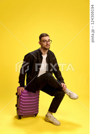 Trendy young man sitting on purple suitcase with confident look, wearing casual black outfit against yellow background. 124309194