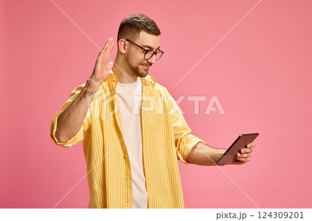 Man in yellow shirt smiling and waving hand while looking at tablet against pink background. 124309201