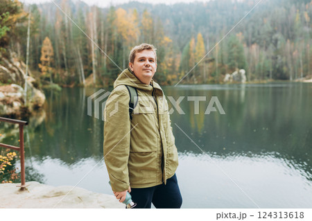 A smiling 30s blonde male traveler with a backpack enjoys the beautiful scenery of the majestic lake. Travel, adventure. Concept of an active lifestyle. A smiling 30s blonde male traveler with a backpack enjoys the beautiful scenery of the majestic lake. Travel, adventure. Concept of an active lifestyle. 124313618